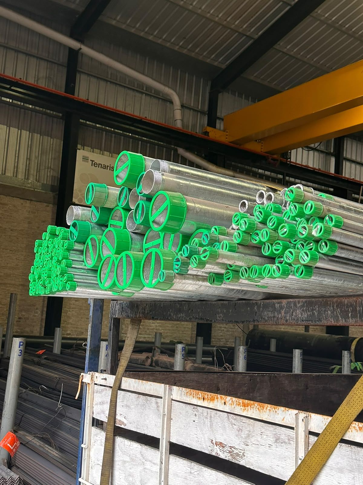 Caños galvanizados con tapas verdes cargados en un camión dentro del depósito, con un puente grúa amarillo.