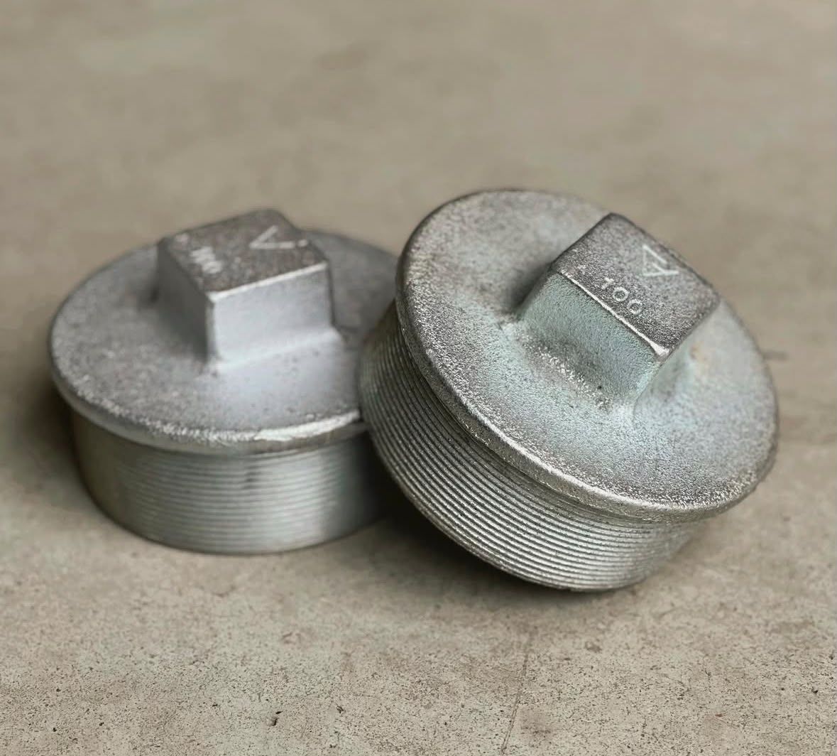 Dos tapones galvanizados con cabeza cuadrada sobre un piso de hormigón.